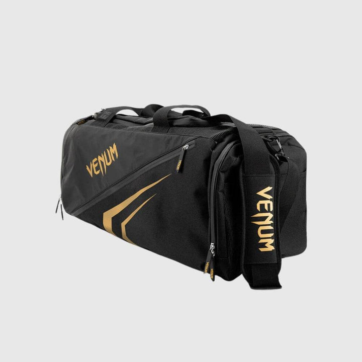 Angled view of the Venum Trainer Lite EVO Sport Bag in black and gold, highlighting the adjustable padded shoulder strap with VENUM branding and zippered side compartments.