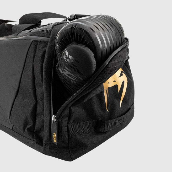 Side compartment of the Venum Trainer Lite EVO Sport Bag unzipped to reveal a pair of black boxing gloves stored inside, with the gold Venum snake logo prominently displayed.