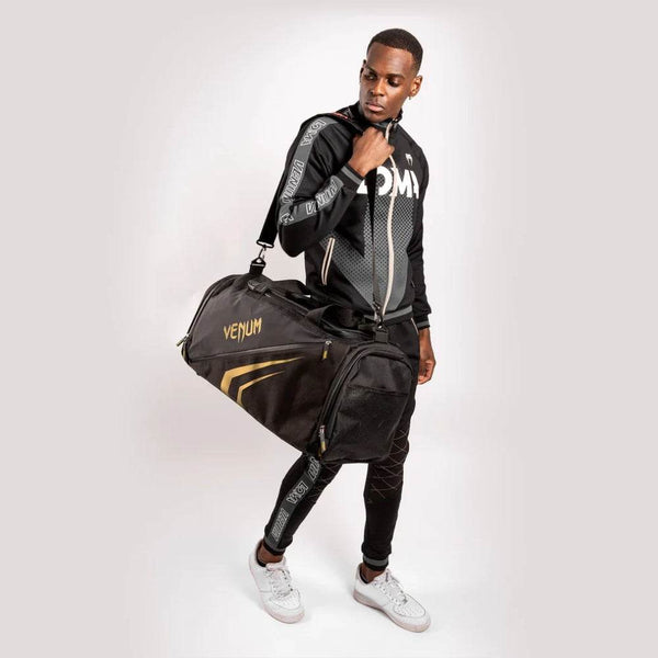 Man dressed in Venum athletic wear carrying a black and gold Venum Trainer Lite EVO Sport Bag over his shoulder, showcasing the bag’s size and stylish design.