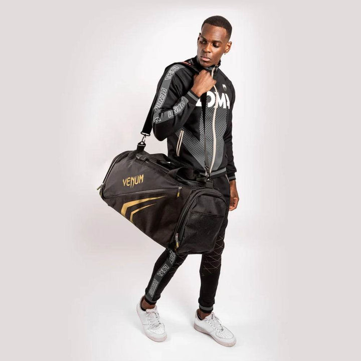 Man dressed in Venum athletic wear carrying a black and gold Venum Trainer Lite EVO Sport Bag over his shoulder, showcasing the bag’s size and stylish design.
