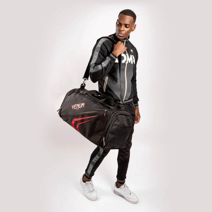 Man carrying the black and red Venum Trainer Lite EVO Sport Bag over his shoulder, wearing Venum athletic gear, highlighting the bag’s stylish contrast design and spacious size.