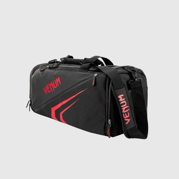 Angled view of the Venum Trainer Lite EVO Sport Bag in black and red, highlighting the bold graphic design, adjustable shoulder strap with VENUM logo, and multiple zippered compartments.