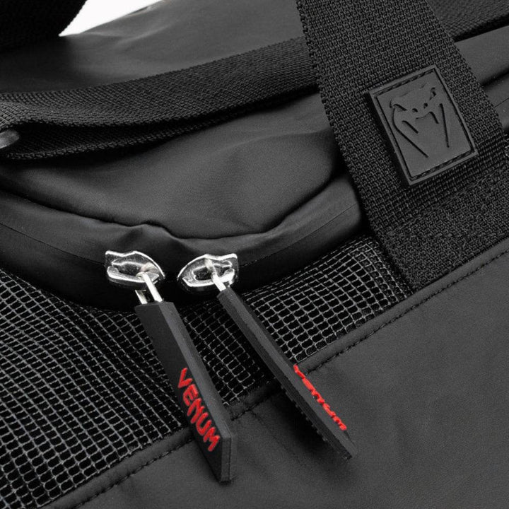 Extreme close-up of the Venum Trainer Lite EVO Sport Bag’s dual zipper pulls with red VENUM branding, mesh ventilation panel, and rubber logo patch stitched to reinforced webbing.