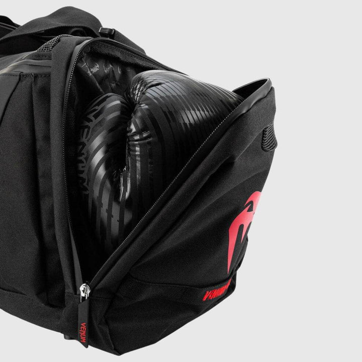 Close-up of the Venum Trainer Lite EVO Sport Bag’s ventilated side compartment unzipped, revealing a pair of black boxing gloves stored inside, with red Venum logo detail on the exterior.