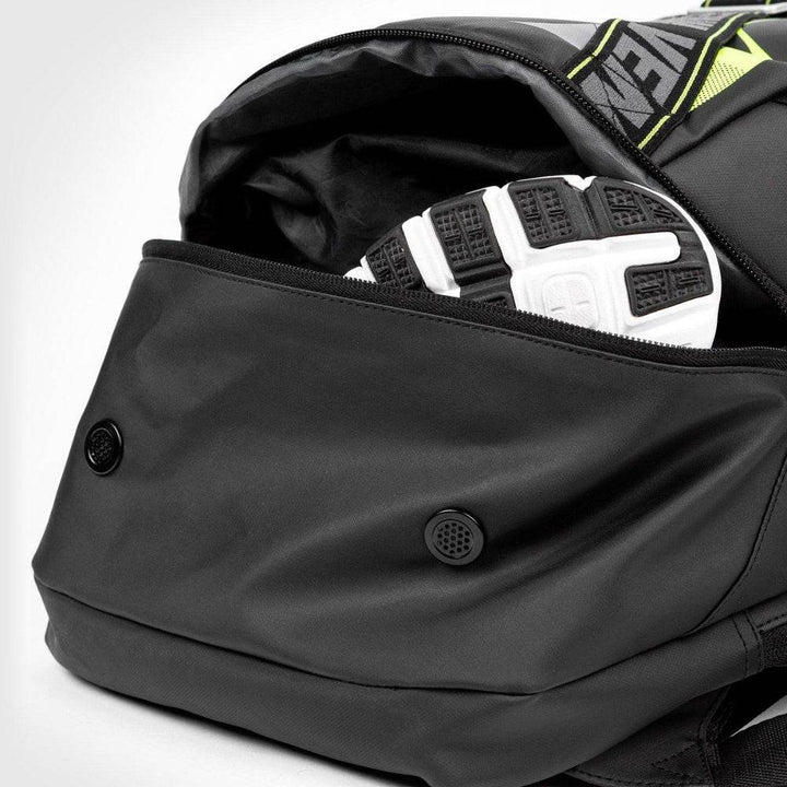 Zippered bottom compartment of the Venum backpack partially open, revealing a pair of athletic shoes inside, with dual ventilation grommets for airflow.