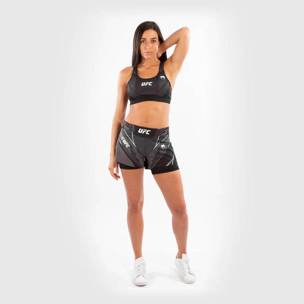 A female athlete poses confidently wearing black UFC Venum women's fightshorts and a matching sports bra. She stands tall with one hand resting behind her head, showcasing the full front fit.