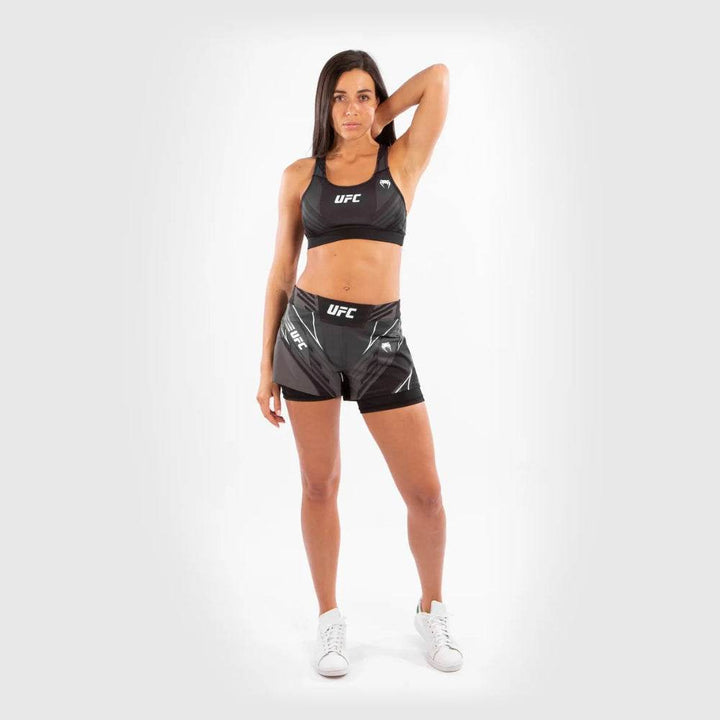 A female athlete poses confidently wearing black UFC Venum women's fightshorts and a matching sports bra. She stands tall with one hand resting behind her head, showcasing the full front fit.